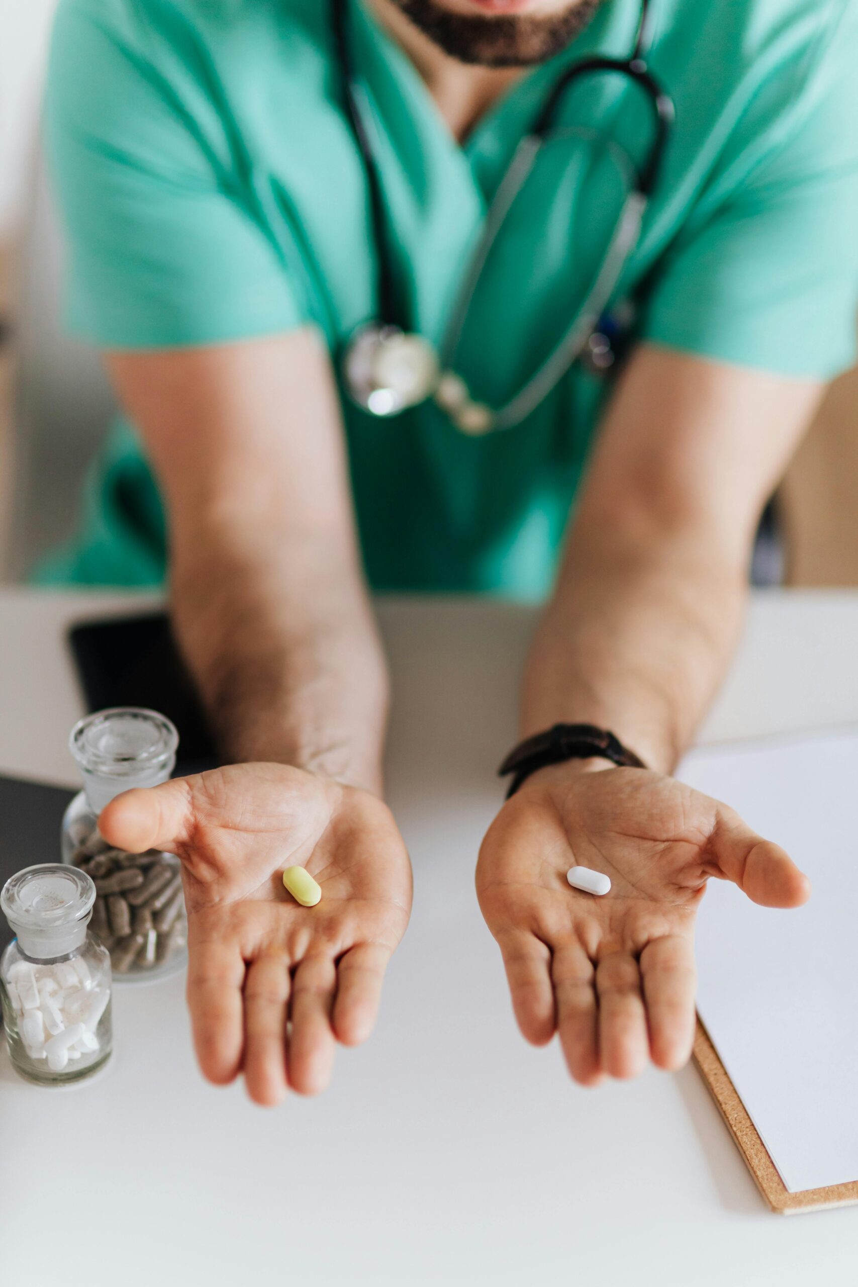 Unrecognizable male doctor wearing medical uniform showing pills to camera while working in modern clinic sitting at table with clipboard and stethoscope